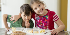Ein Bub und ein Mädchen sitzen am Tisch, lachen und essen Apfelstücke, Schüssel mit Kichererbsen steht vor ihnen
