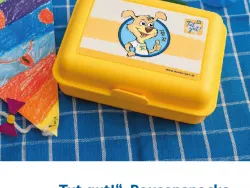 gelbe Jausenbox auf blau kariertem Geschirrtuch, links davon ein bunter Papierdrache