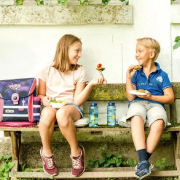 Mädchen und Bub sitzen auf Bank und jausnen.